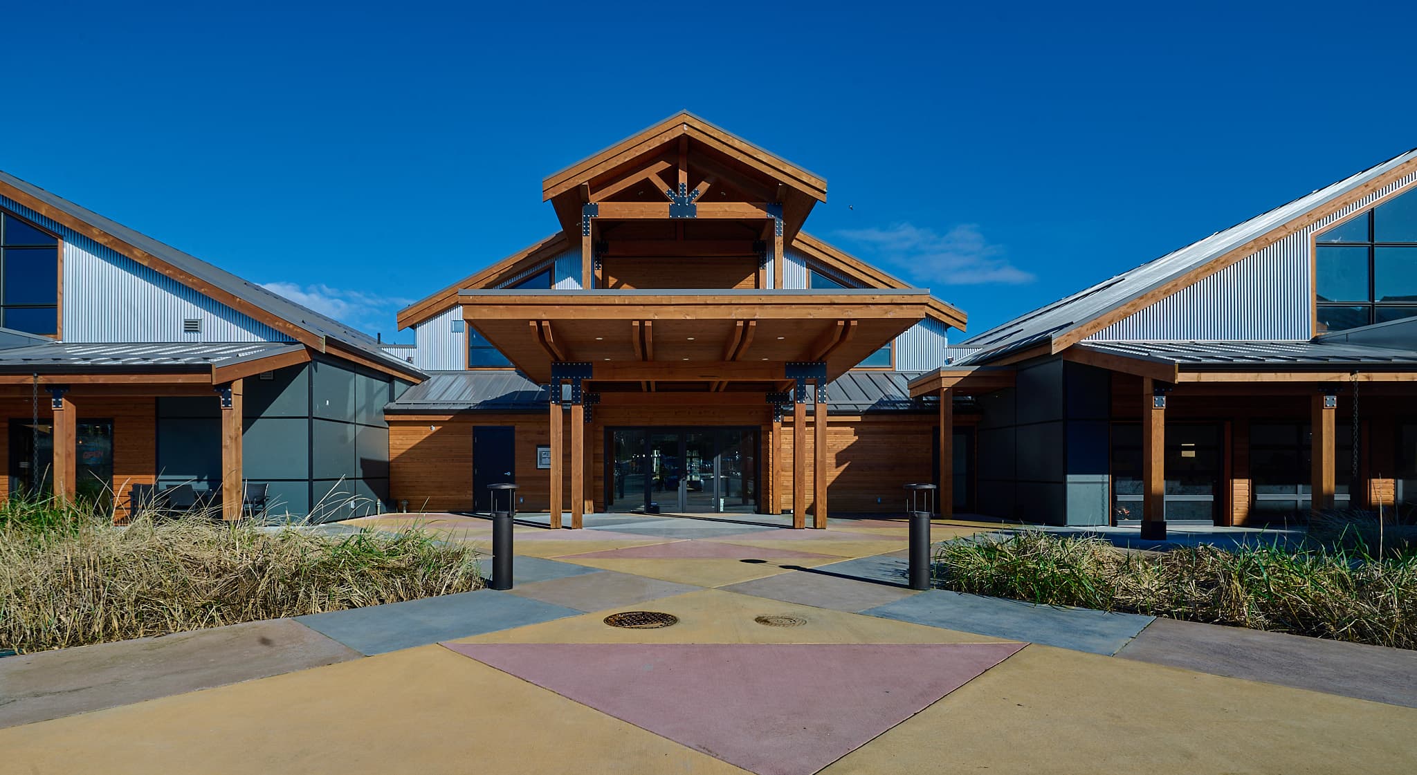 Modern rustic building with a large wooden entrance and a colorful geometric patterned concrete plaza.