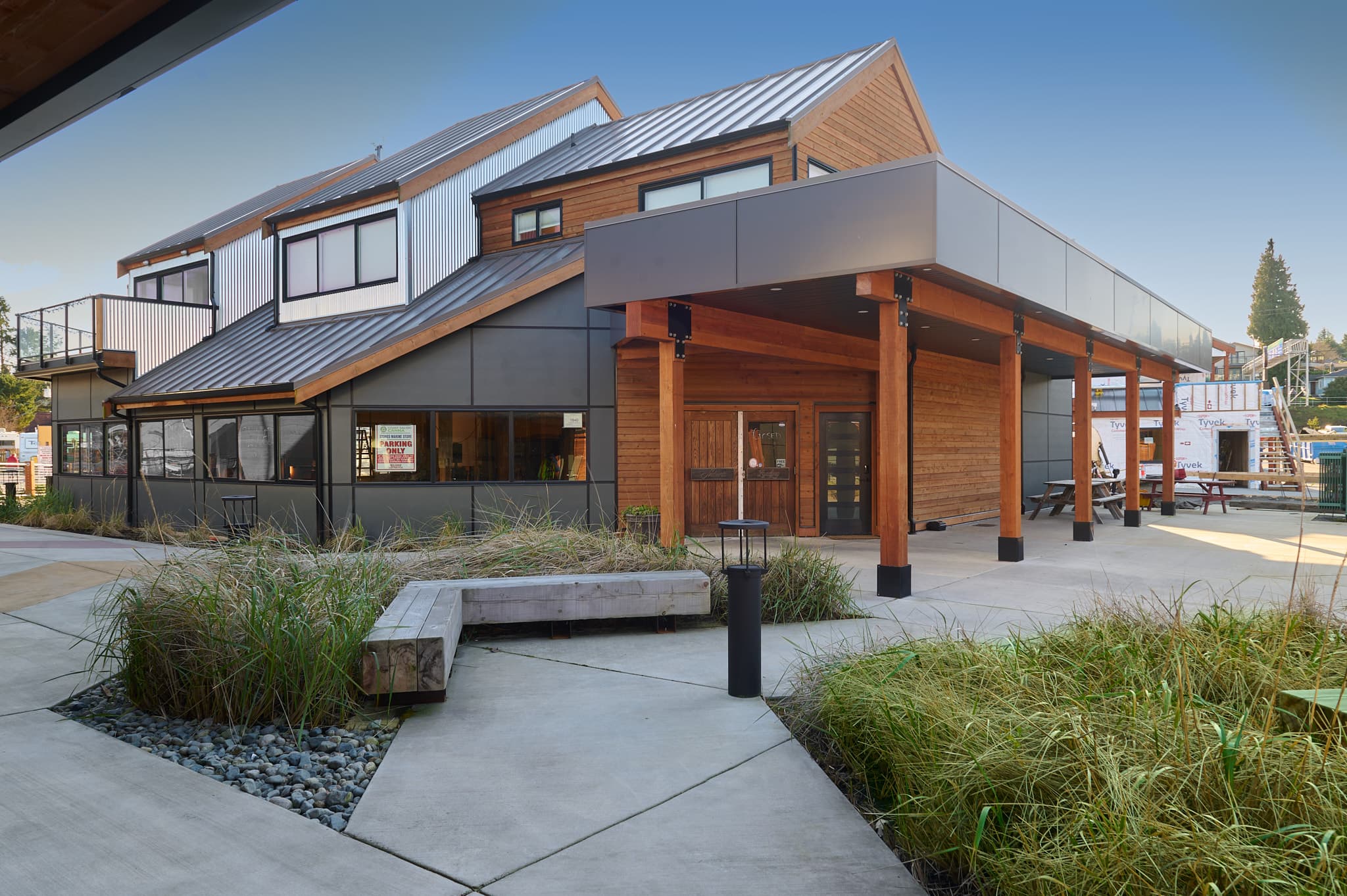 Modern multi-story building with wood and metal siding, timber overhang, and landscaped concrete paths.