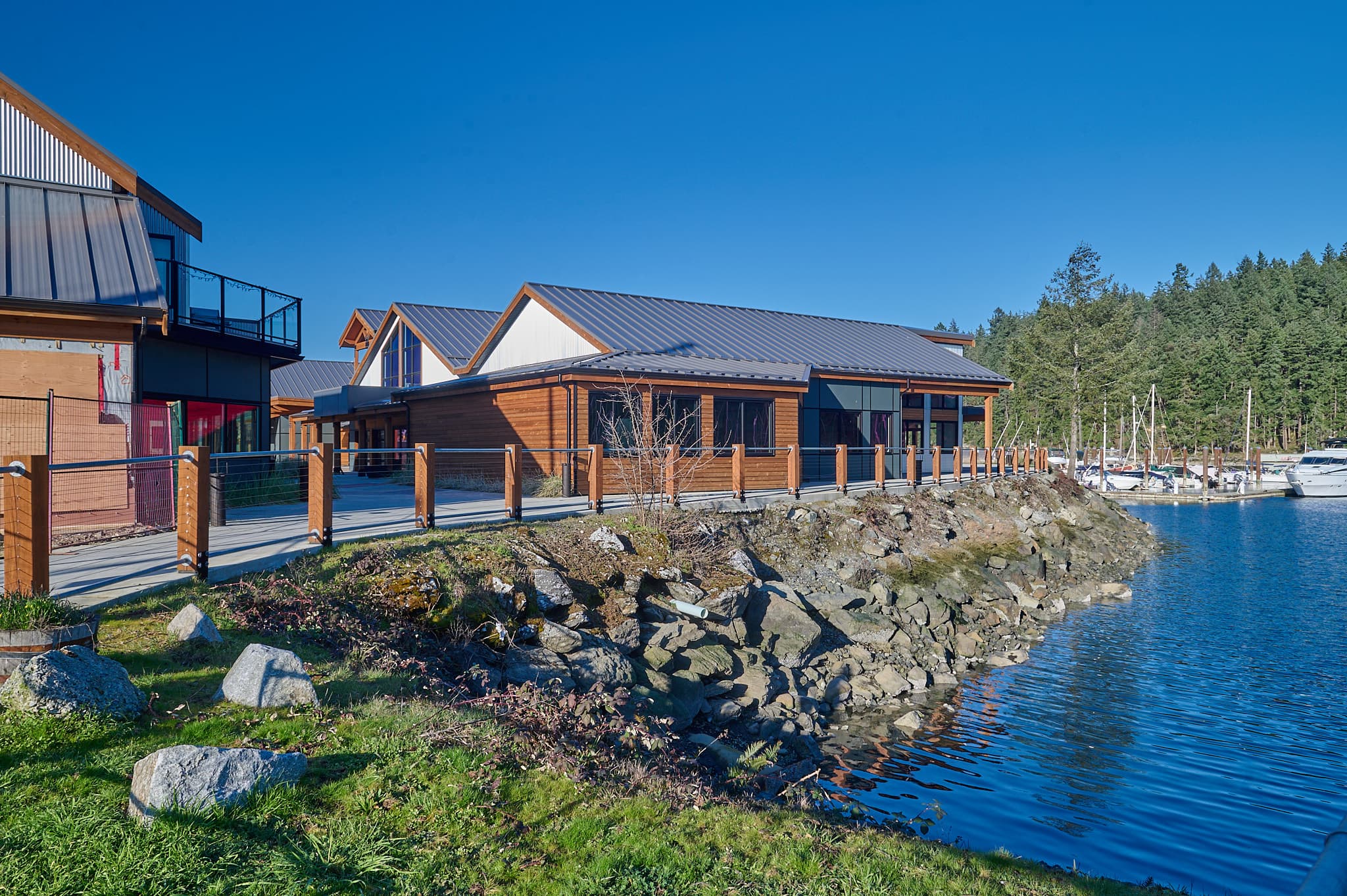 Modern wooden buildings along a rocky shoreline overlooking a marina and dense green forest.