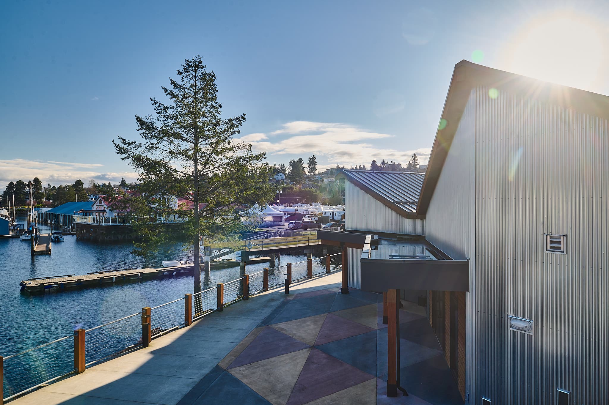 Modern building and geometric walkway beside a sunlit marina with docks and evergreen trees.