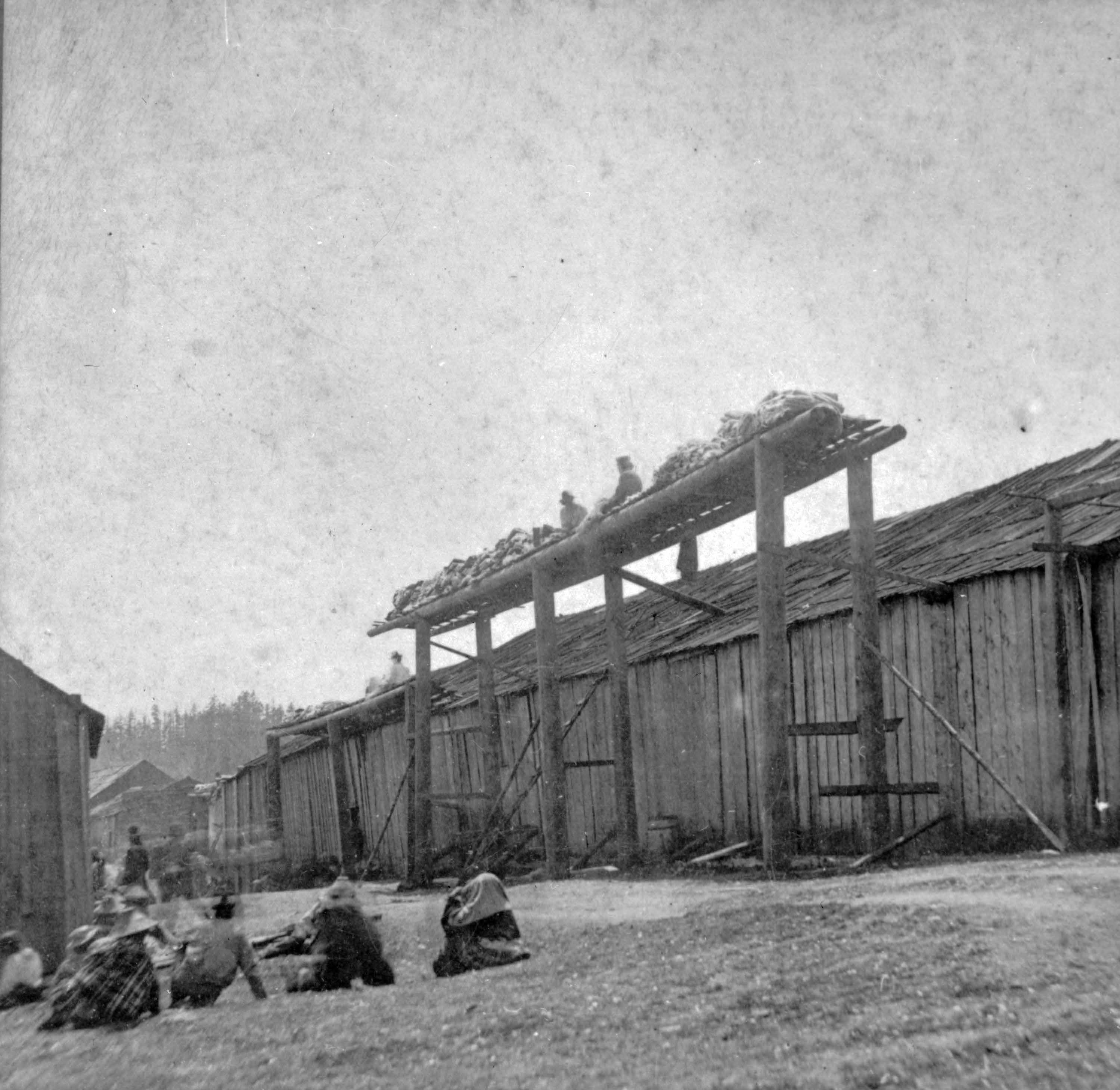 People sitting on the ground and atop a high wooden rack outside a longhouse.